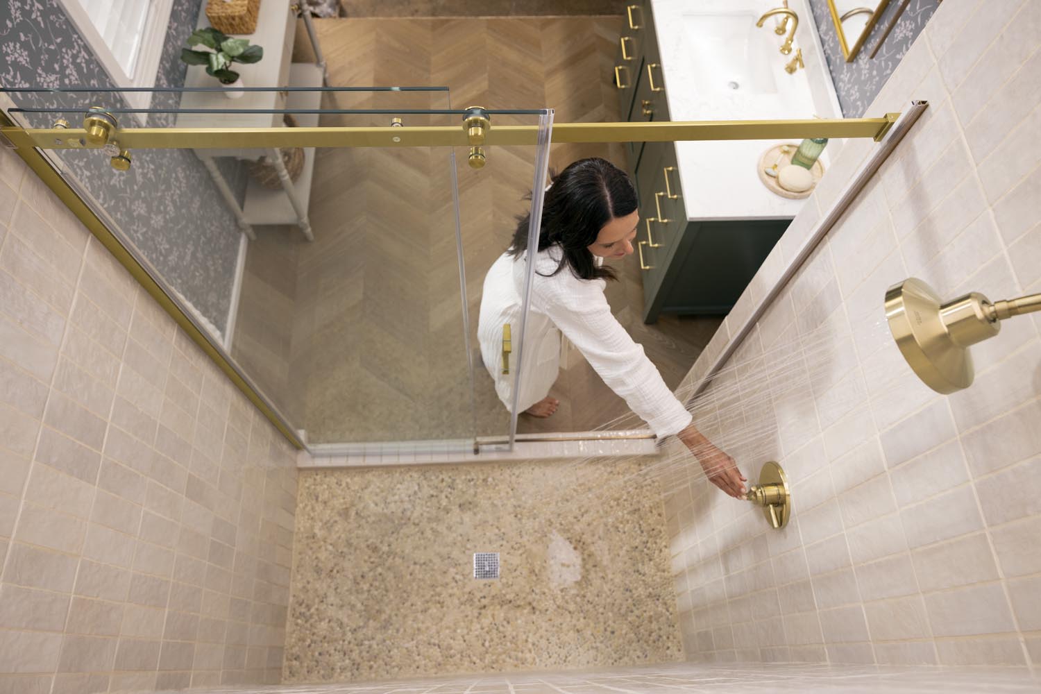 Person opening a glass shower door in a bathroom with gold fixtures.